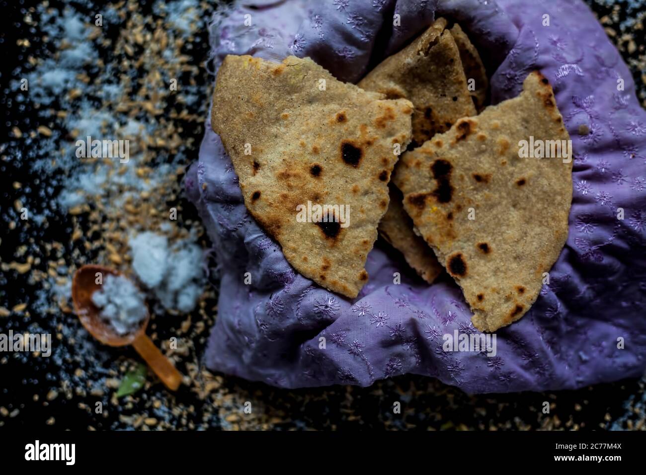 Nahaufnahme von runden Brot Bhakri auf der schwarzen Holzoberfläche zusammen mit etwas rohem Vollkorn, Salz in einem Behälter. Schuss von Bhakri in einem Container auf Stockfoto