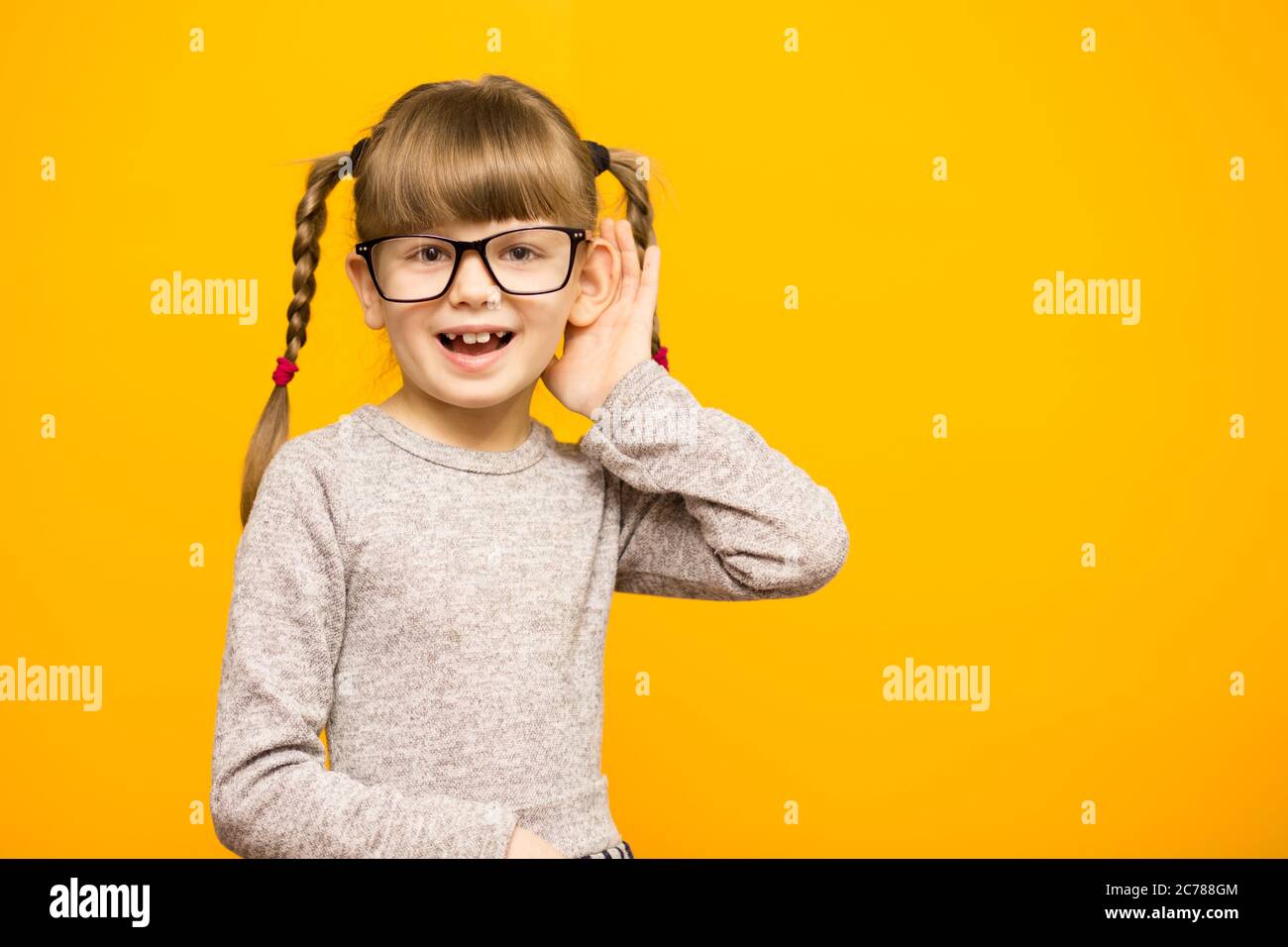 Kleines Mädchen Journalistin in Brille mit überrascht euphoria Gesicht und lustige Zöpfe hören etwas hält seine Hand an Ohr isoliert auf yel Stockfoto