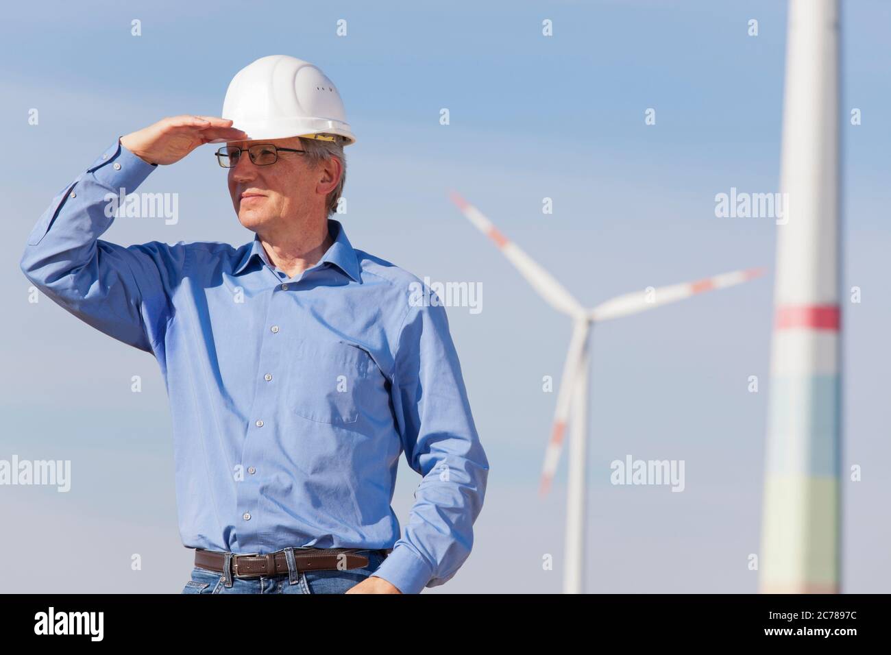 Ingenieur mit Hut vor einem Windpark auf der Suche nach einer strahlenden Zukunft für grüne Energie - konzentrieren Sie sich auf die Person Stockfoto