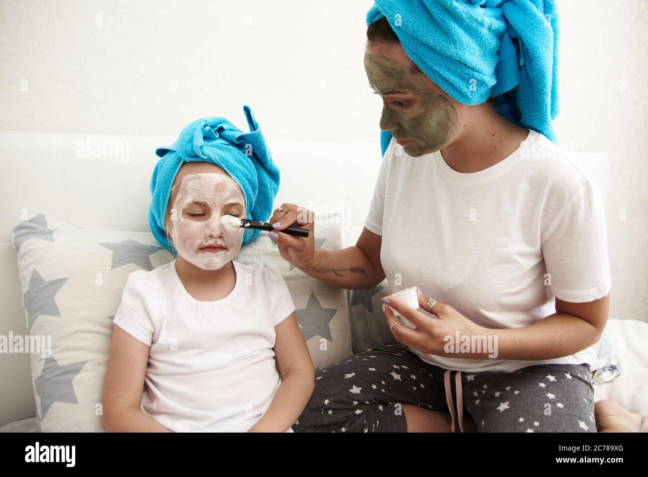 Nette junge Mutter und ihre Tochter sitzen auf dem Bett in Handtüchern auf ihrem Kopf und machen eine Gesichtsmaske. Hautpflege, Familienbeziehungen. Stockfoto