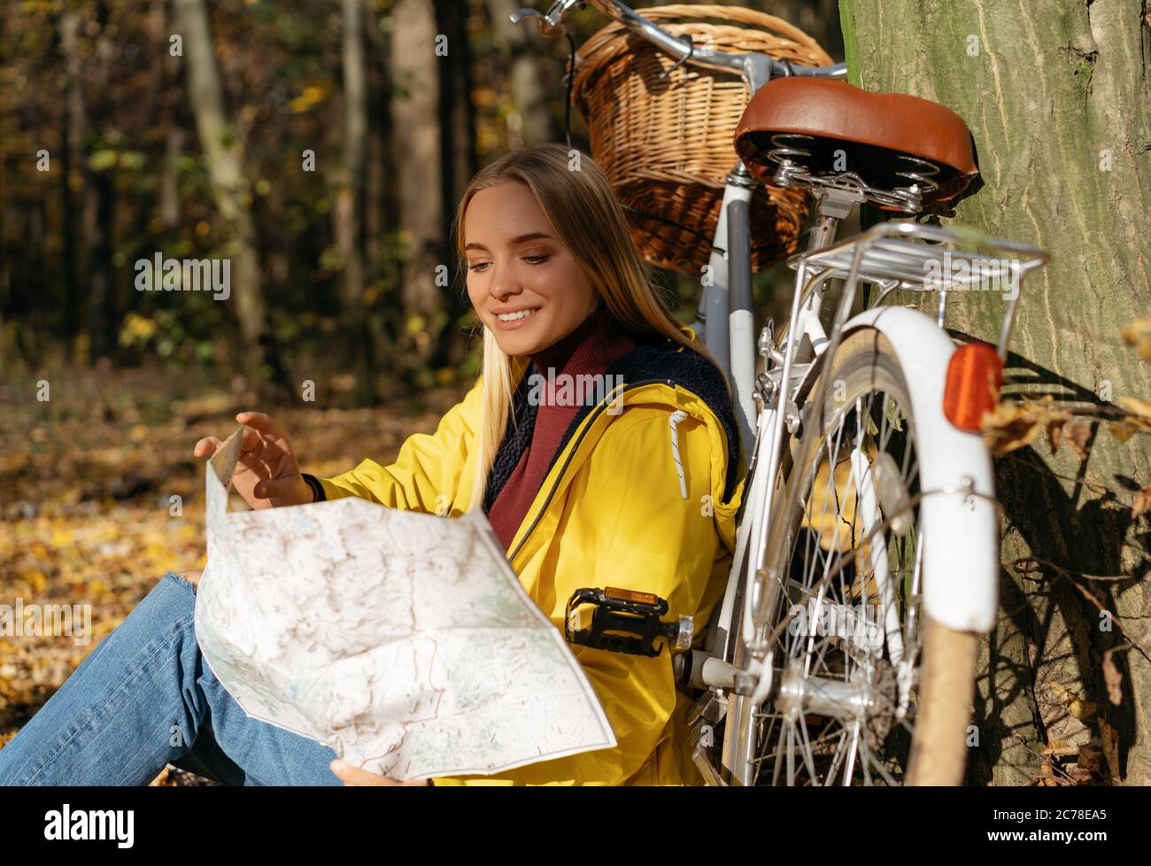 Schöne lächelnde Frau in gelbem Regenmantel mit Reisekarte, die in der Nähe des Fahrrads im Wald sitzt. Reisekonzept Stockfoto