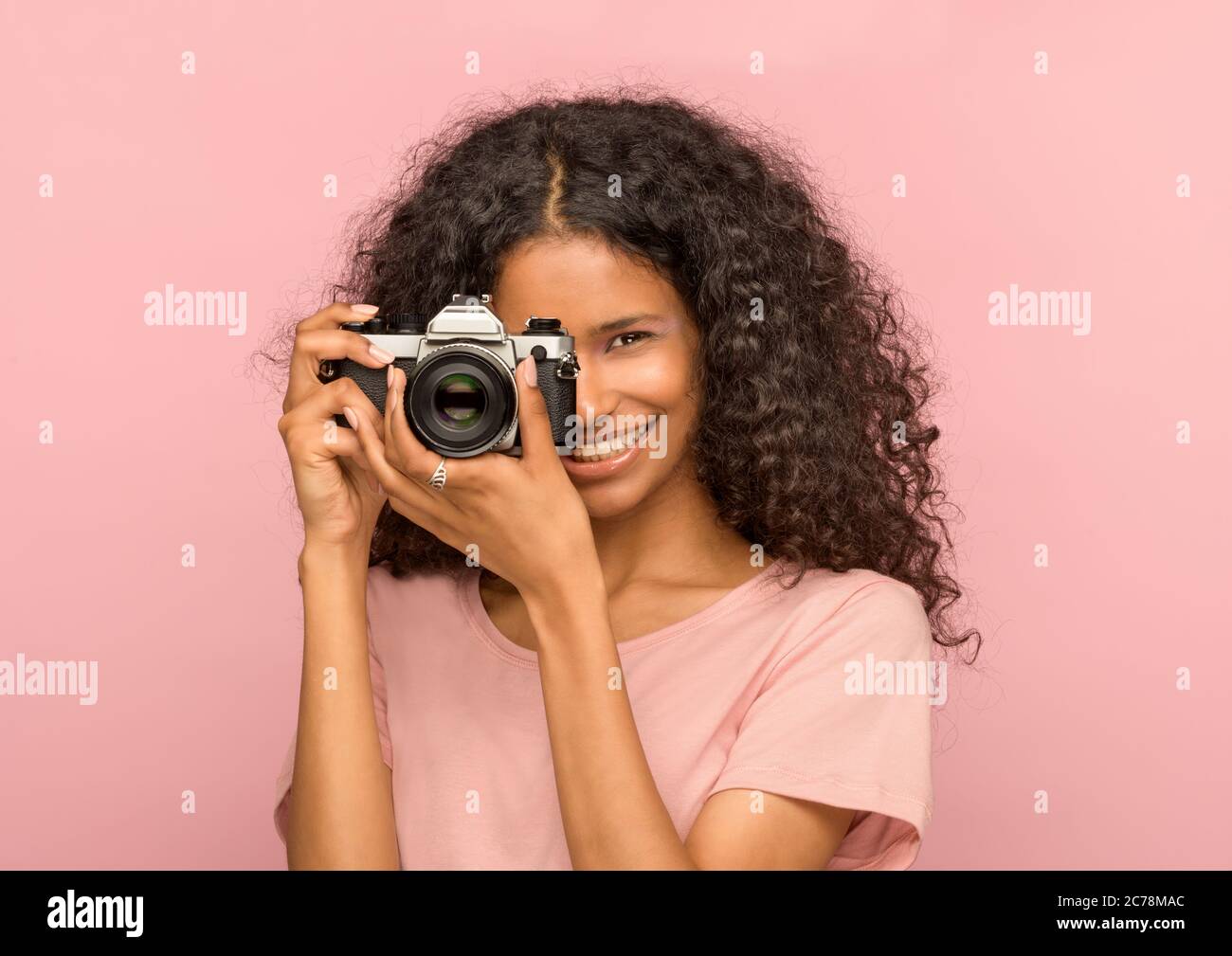 Glücklich attraktive junge schwarze Frau mit einer Vintage-Kamera konzentriert sich auf den Betrachter mit einem strahlend freundlichen Lächeln vor einem rosa Studio Hintergrund Stockfoto
