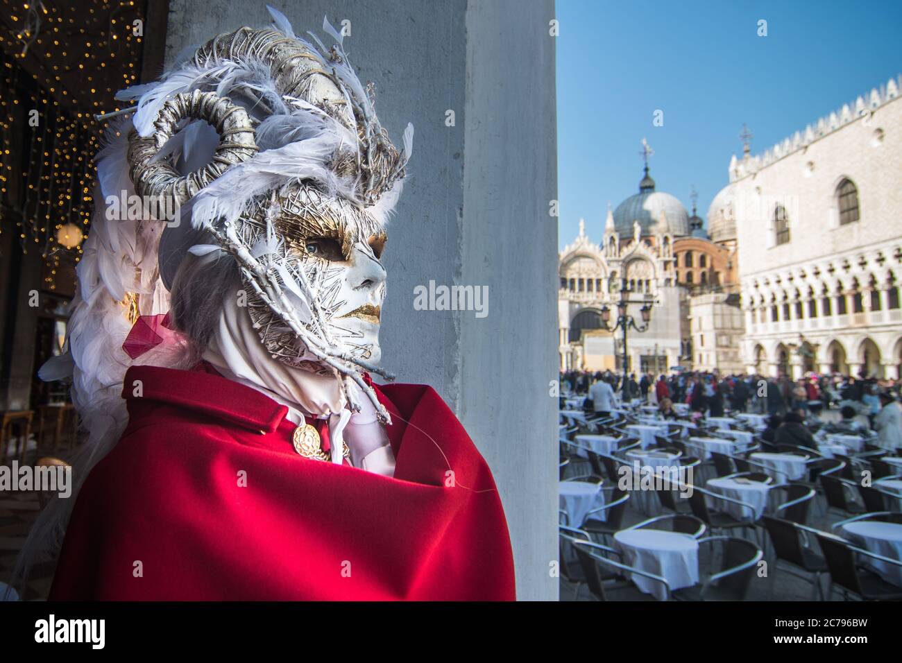 VENEDIG, ITALIEN - 28. FEBRUAR 2019: Eine elegante weiße Maske mit rotem Umhang in der Nähe einer Reihe von Bartischen während des Karnevals in Venedig Stockfoto