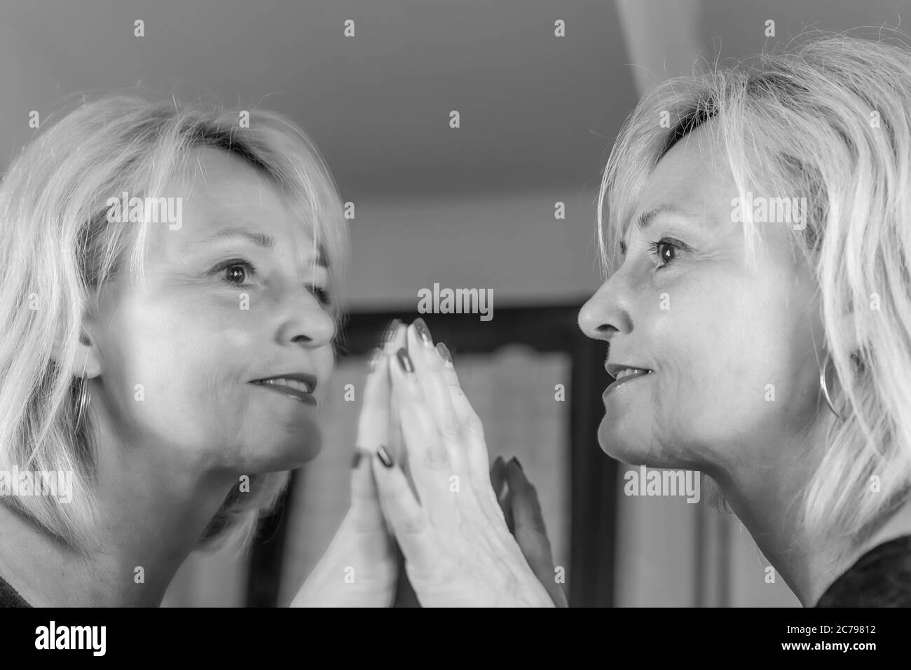 Eine blonde Frau mittleren Alters spiegelt sich in einem großen Spiegel wider, indem sie ihn mit ihren Händen mit einem glücklichen Ausdruck berührt Stockfoto