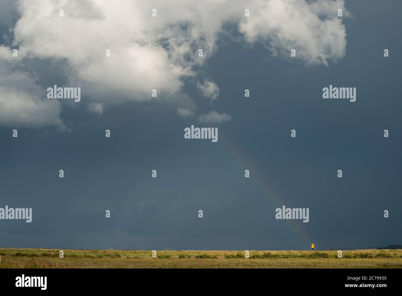 Einfaches, aber grafisches Bild einer einzigen Figur in gelber Kleidung, die am Fuße eines Regenbogens gegen einen dunklen Himmel und eine große weiße Wolke steht Stockfoto