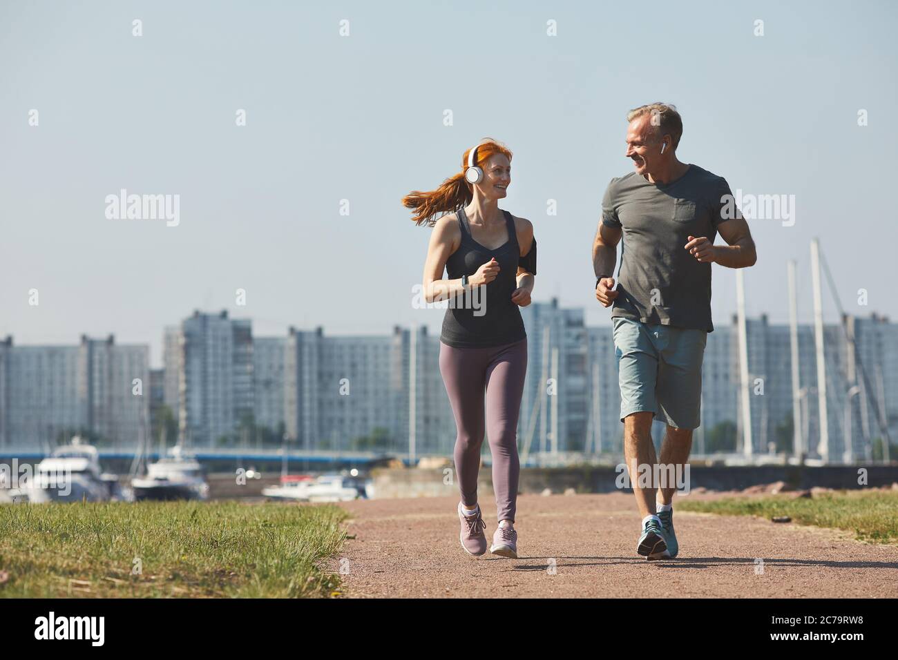 Sportlich reifes Paar in Kopfhörern lächelt sich an, während sie gemeinsam in der Stadt joggen Stockfoto