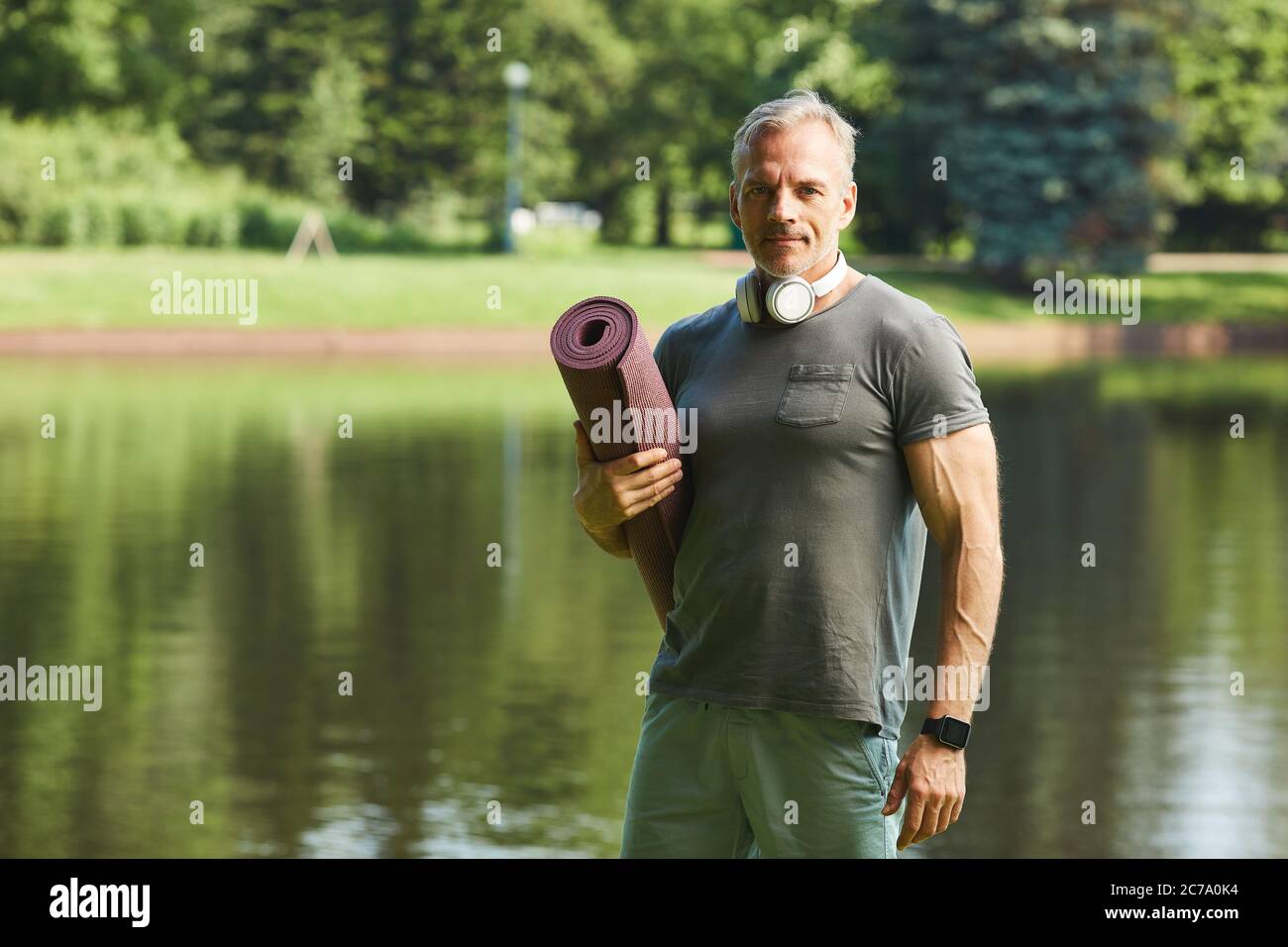Portrait of content muskulöser reifer Mann mit Kopfhörern um den Hals hält Übungsmatte gegen Park See Stockfoto