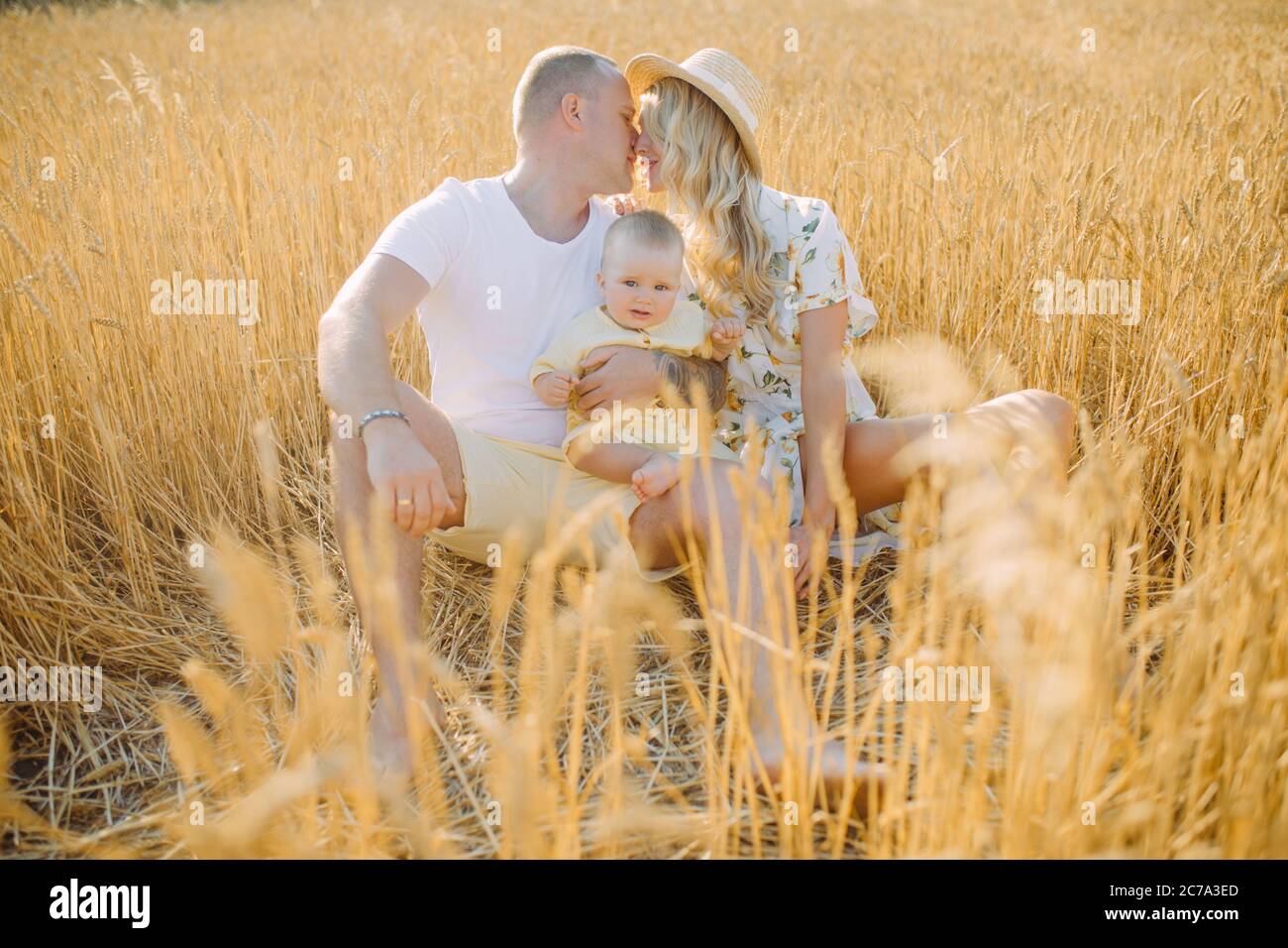 Glückliche junge Eltern mit Baby ruht zwischen gelben Weizenfeld und sie küsst. Stockfoto