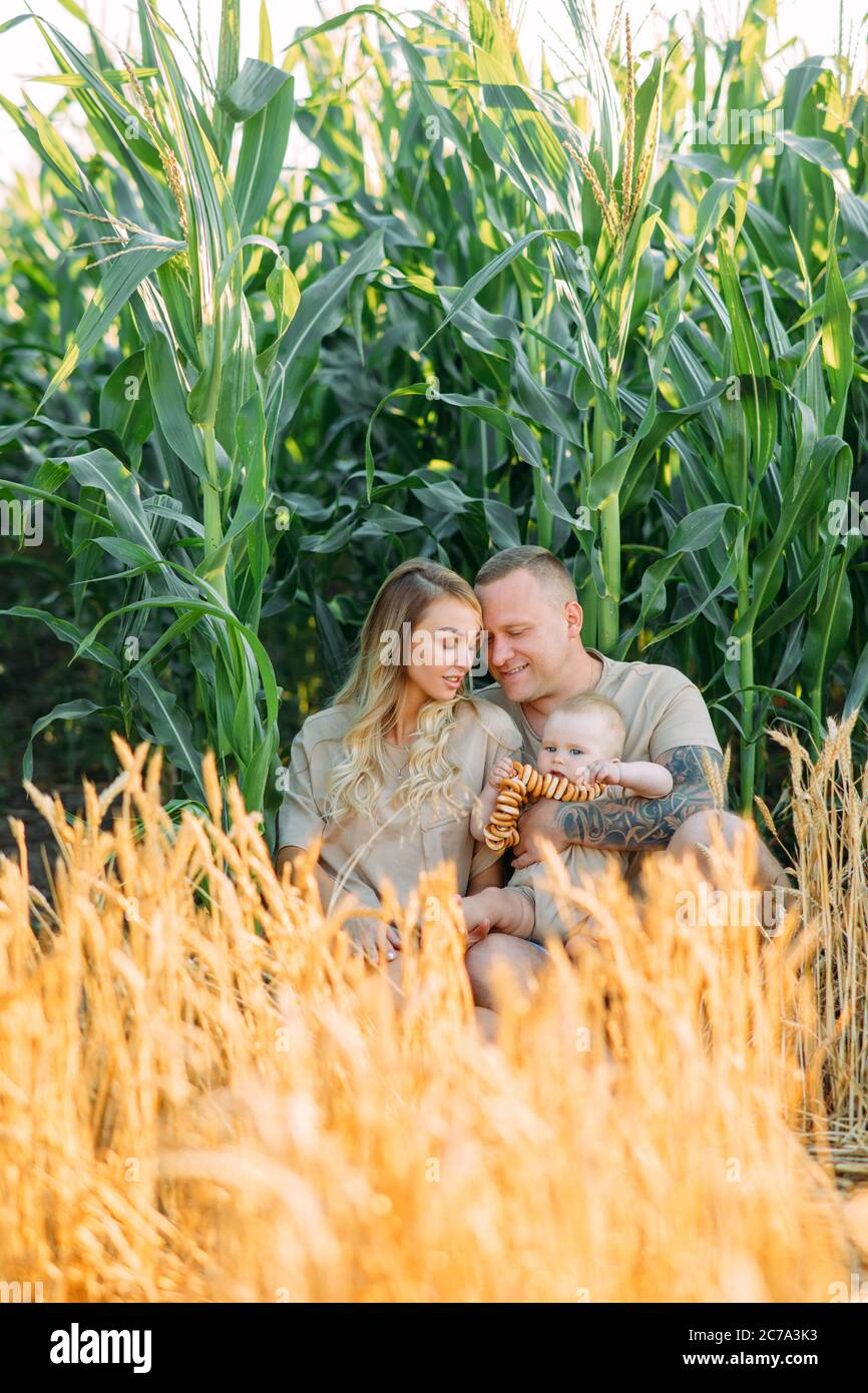 Glückliche junge Familie mit Baby sitzt und ruht unter gelbem Weizenfeld in der Nähe von Mais. Stockfoto