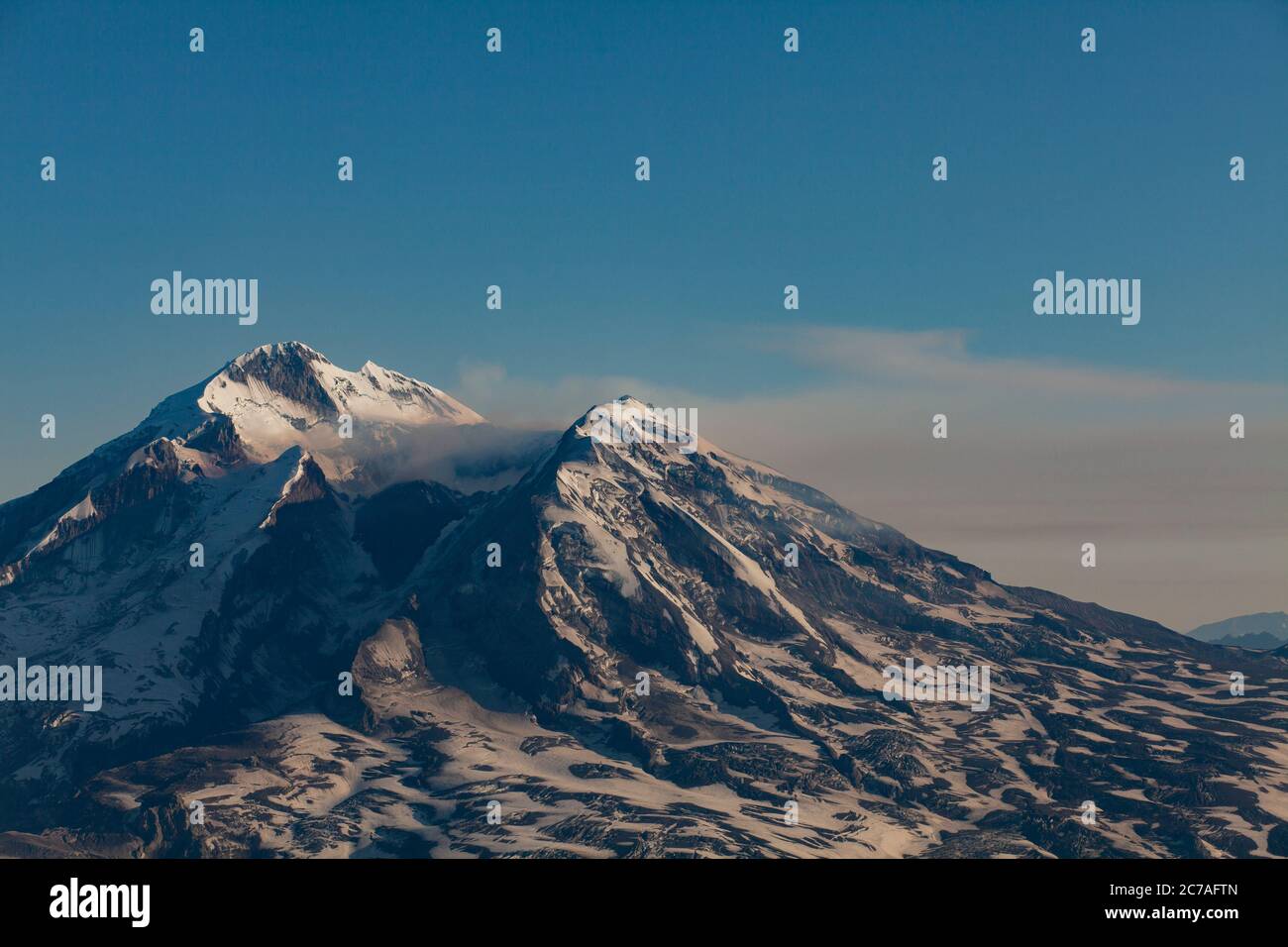 Schneebedeckter vulkanischer Berg mit einer Rauchwolke, die über den Himmel strömt und die dramatische und zerklüftete Wildnis Alaskas zeigt. Stockfoto