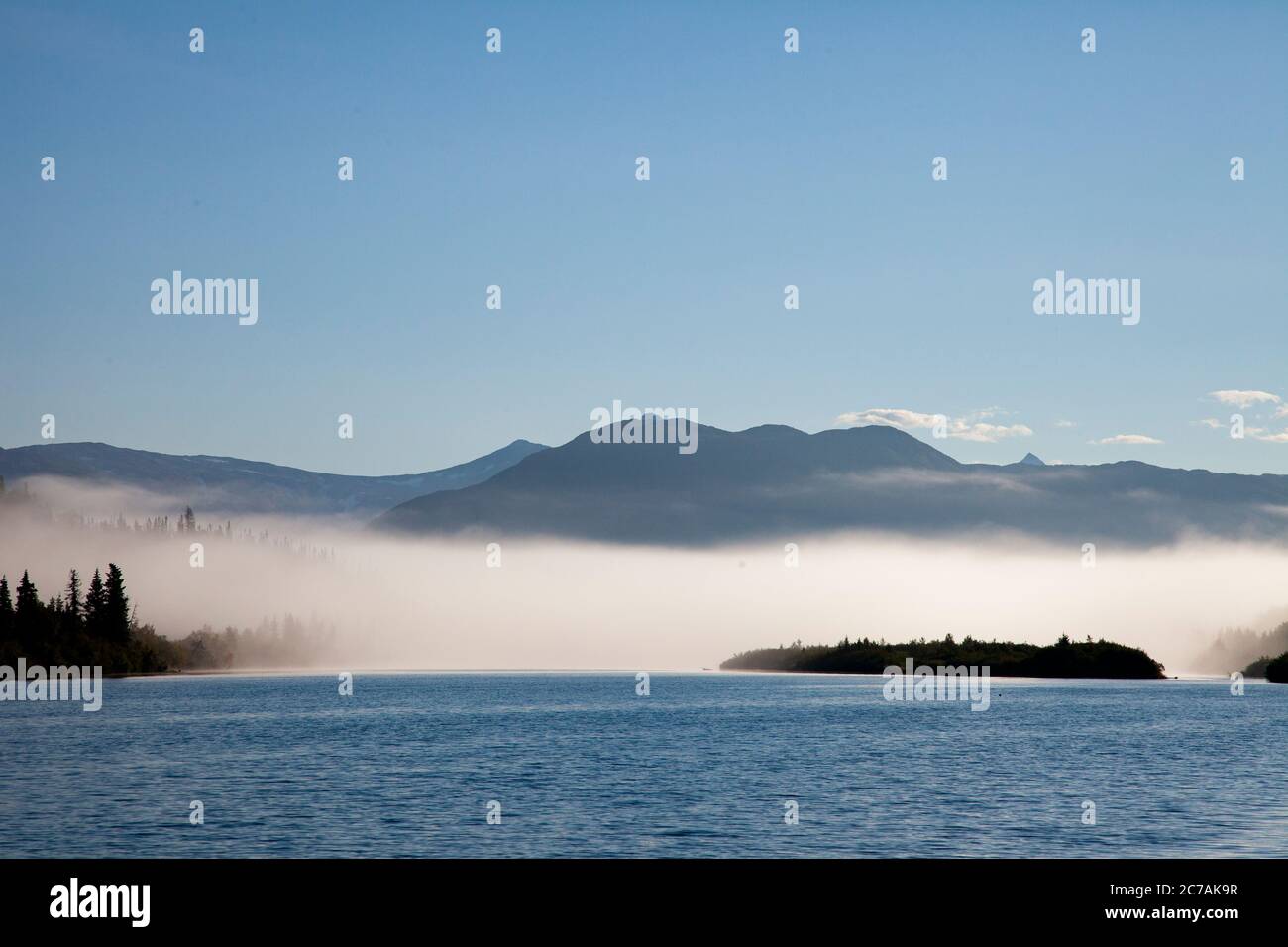 Der Morgennebel zieht über den Lake Iliamna, Alaska, mit Bergen im Hintergrund und Sonnenlicht, das sich vom ruhigen, ruhigen Wasser der Wildnis reflektiert Stockfoto