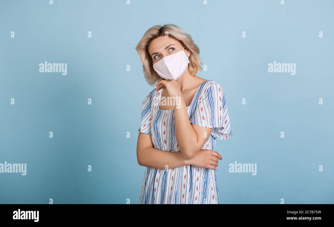 Nachdenkliche kaukasische Frau mit blonden Haaren, die ihr Kinn berühren und in einem Kleid an einer blauen Wand im Studio posieren Stockfoto