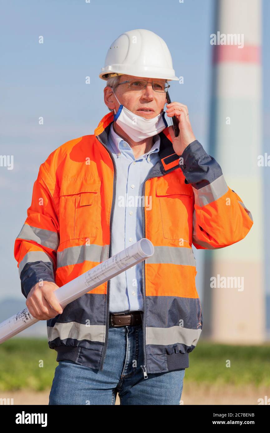 Ingenieur oder Architekt, der eine Gesichtsmaske aus Coronavirus und Blaupause in der Hand auf einer Baustelle trägt, die am Handy spricht Stockfoto