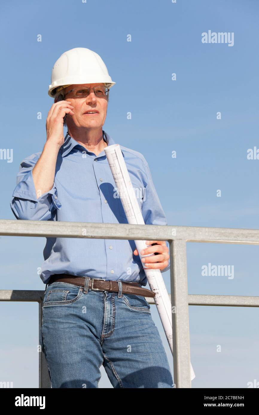 Ingenieur oder Architekt mit Hut und Blaupause in der Hand auf einer Baustelle, die am Handy spricht Stockfoto