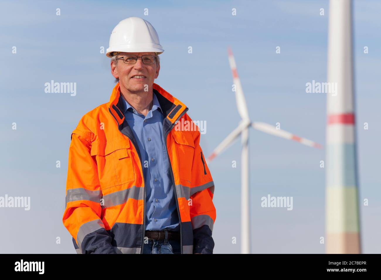 Lächelnder Ingenieur mit Hut und Schutzkleidung an einem sonnigen Tag vor Windmühlen Stockfoto