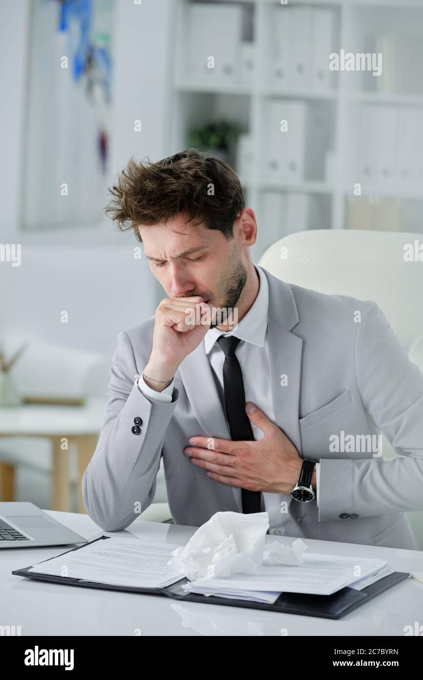 Schlecht aussehender Geschäftsmann mit Bart, der mit Dokumenten am Schreibtisch sitzt und die Hand auf der Brust hält, während er Husten, Coronavirus oder Lungenentzündung hat Stockfoto