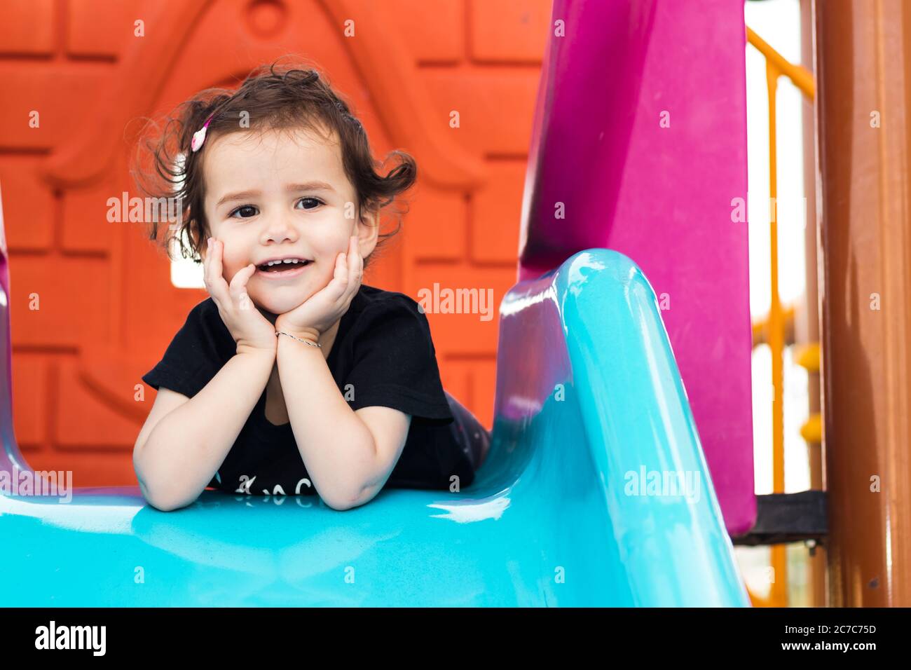 Ein süßes kleines Mädchen, das auf der blauen Rutsche auf einem Spielplatz mit Händen unter dem Kinn liegt. Stockfoto