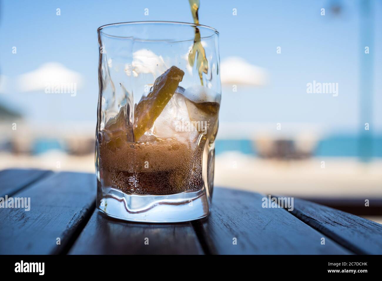 Ein Softdrink wird in ein Glas mit Eis und Zitrone gegossen, im Hintergrund sieht man Sonnenschirme, Himmel und Strand. Stockfoto