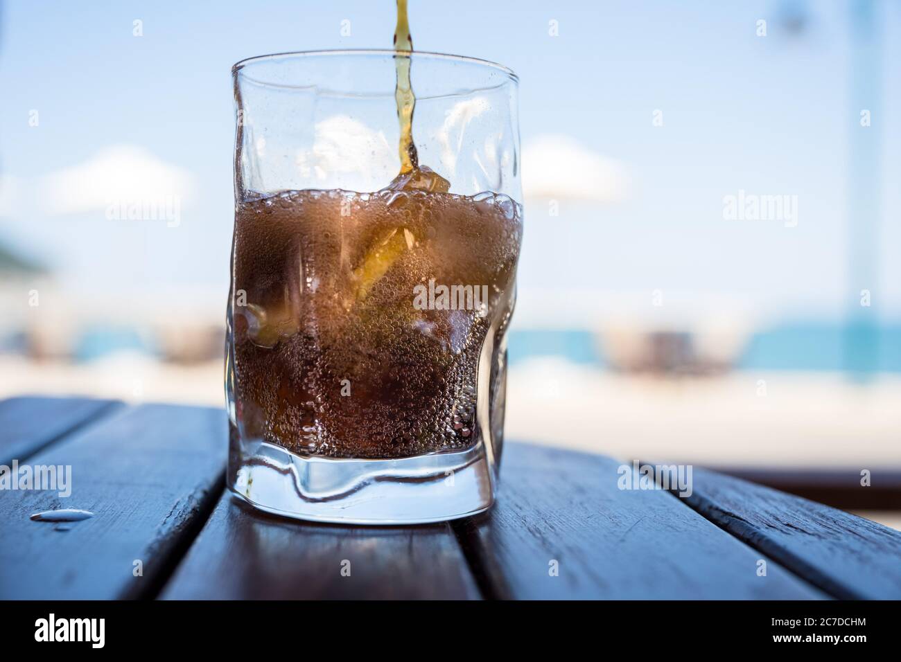 Ein Softdrink wird in ein Glas mit Eis und Zitrone gegossen, im Hintergrund sieht man Sonnenschirme, Himmel und Strand. Stockfoto