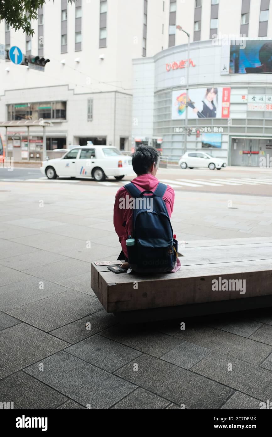 MATSUYAMA, JAPAN - 21. Sep 2019: Eine vertikale selektive Aufnahme einer Person mit schwarzem Rucksack, die auf einer Bank auf dem Bürgersteig in der Nähe von Autos und Baustellen sitzt Stockfoto