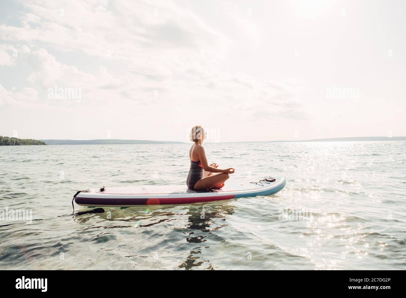 Frau aus dem mittleren Alter, die bei Sonnenuntergang Yoga auf dem Paddle sup Surfbrett praktiziert. Weibliche Stretching macht Training auf See Wasser. Moderner Individualsport Stockfoto