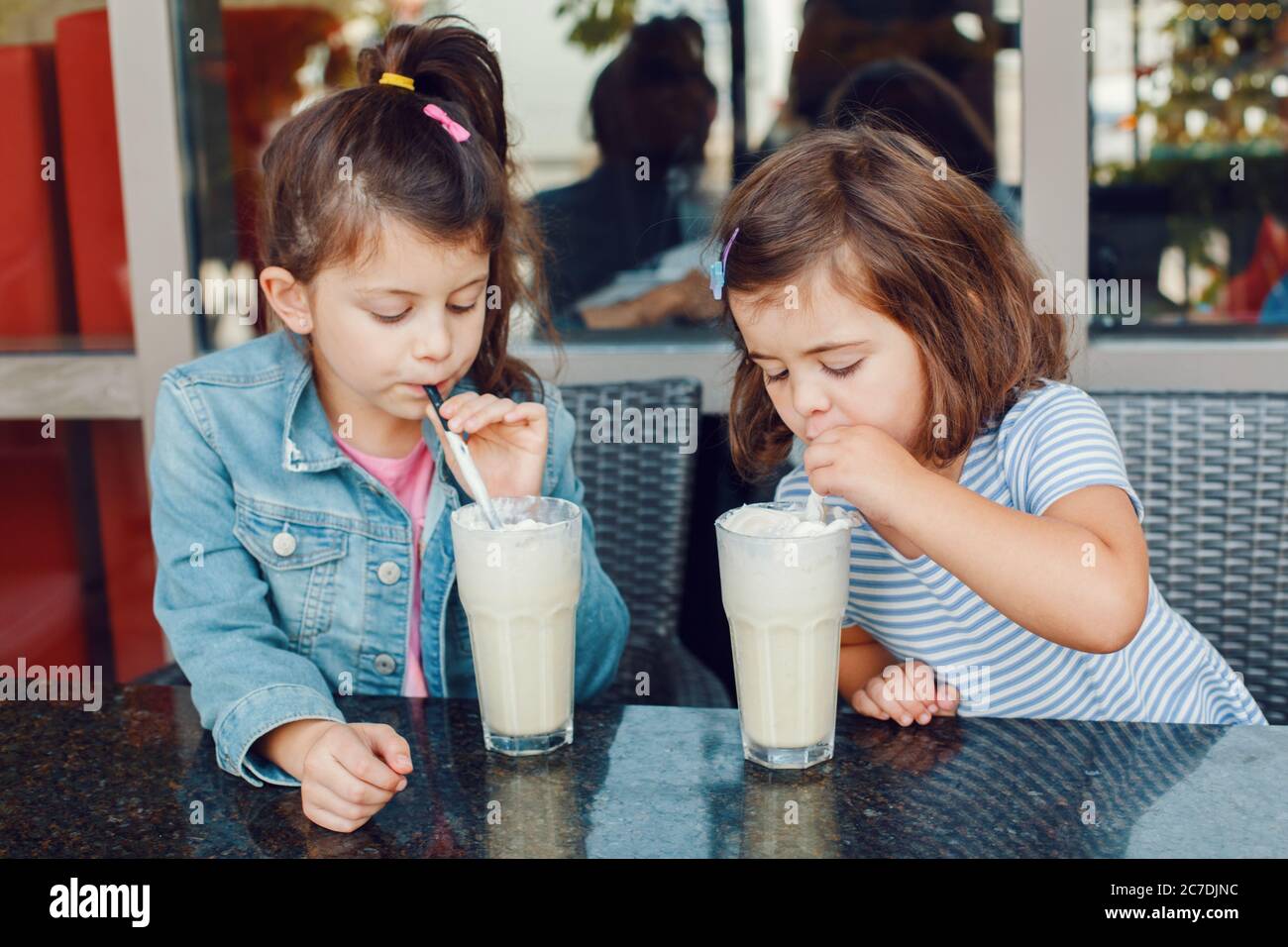 Zwei lustige Schwestern der kaukasischen Vorschule trinken im Café Schluck Milchshakes. Freunde Mädchen, die ein Brunch-Frühstück im Freien essen. Kalte Sommerdessspeisen Stockfoto