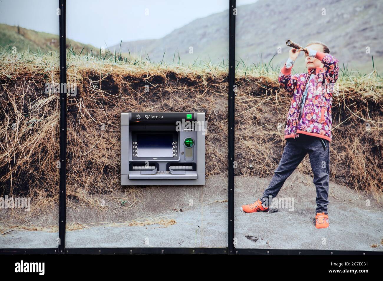 Geldautomat auf dem Bankgebäude mit einem riesigen Foto eines jungen Mädchens, das Teleskop in der Wildnis benutzt. Miðvágur.Vagar.Färöer-Inseln.Territorium von Dänemark Stockfoto