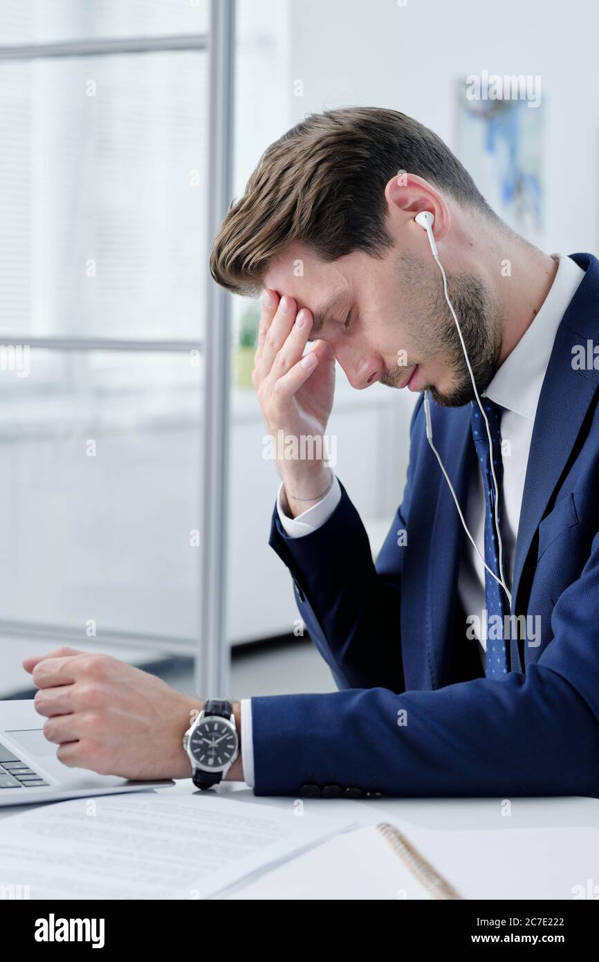 Junger Geschäftsmann in Ohrhörern müde von der Büroarbeit sitzen am Schreibtisch und reiben Stirn, während das Gefühl Kopfschmerzen Stockfoto
