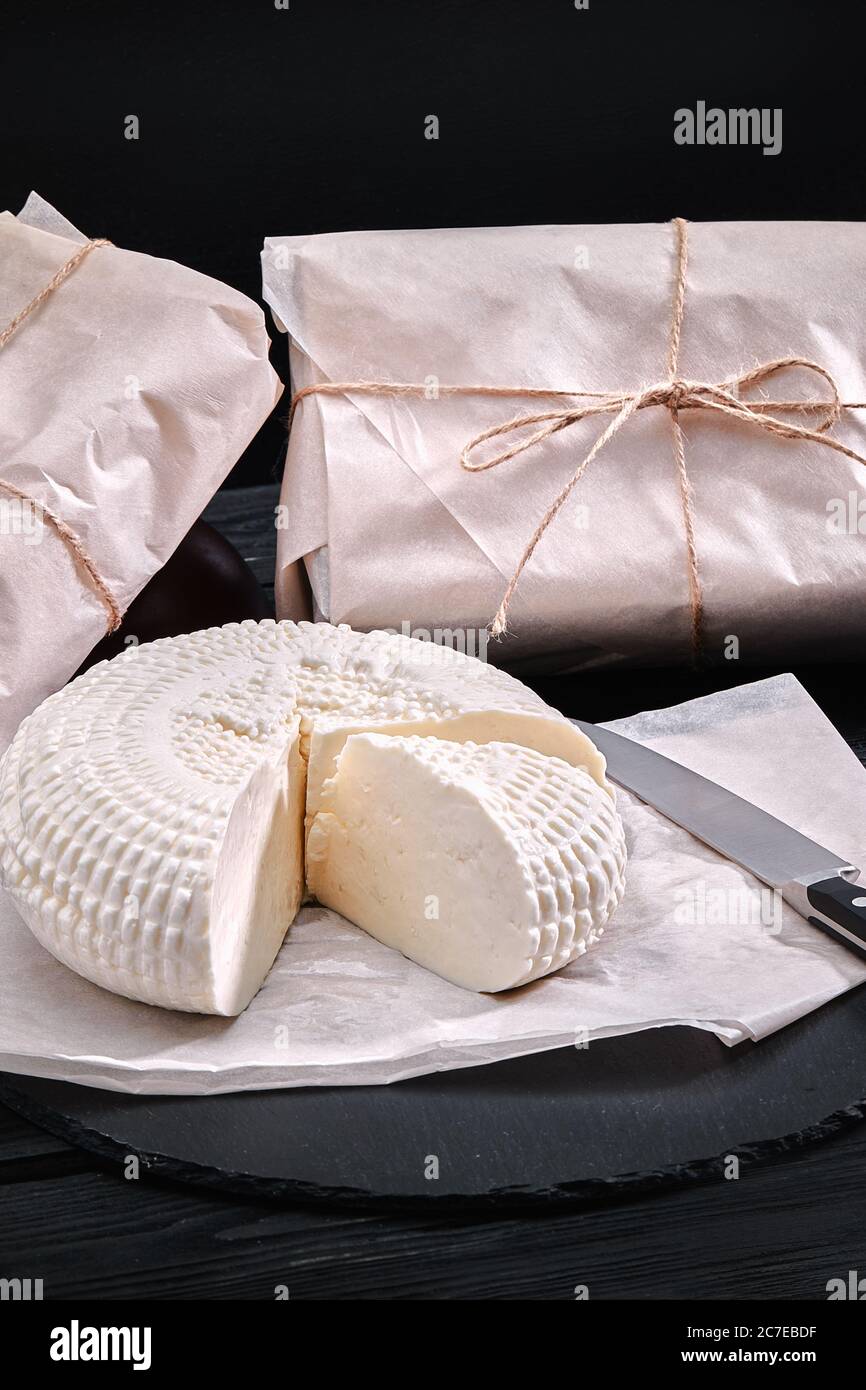 Adyghe Käse Schnitt ein dreieckiges Stück ab. In der Nähe befinden sich drei Käseköpfe, die in Papier verpackt sind. Auf einem Holzbrett. Hintergrund grauer Beton Stockfoto