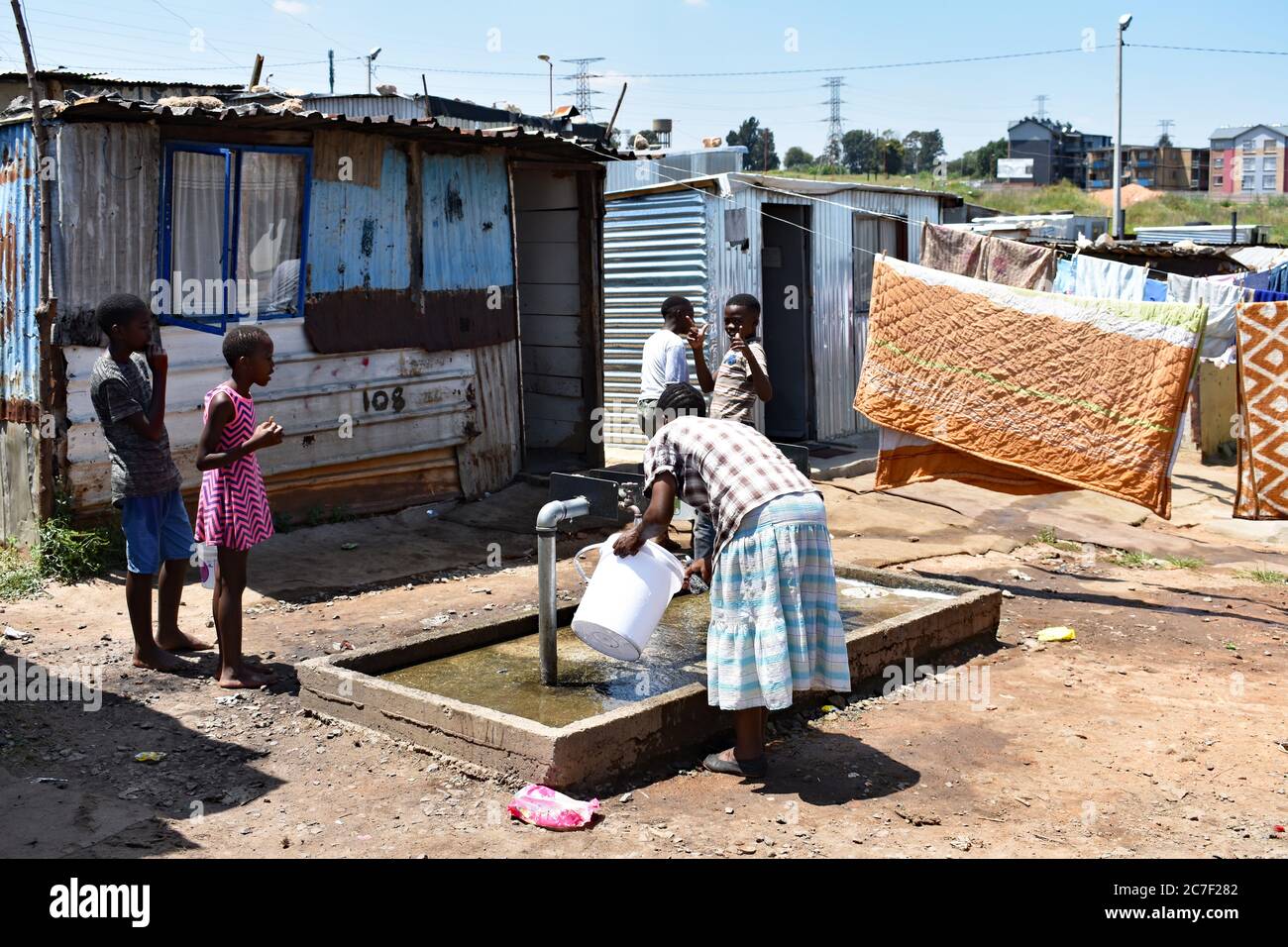 Eine Mutter füllt einen Eimer Wasser aus einem Wasserhahn, während ihre Kinder in einem Slum in Soweto, Johannesburg, auf sie schauen. Kleidung und Bettwäsche trocknen auf der Wäscheleine Stockfoto