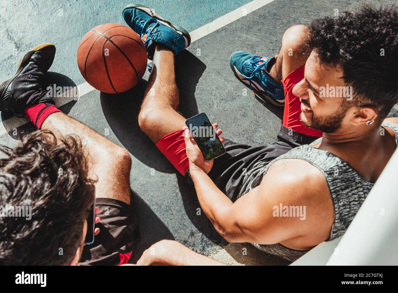 Der junge Mann ruht nach dem Basketballspiel und nutzt sein Smartphone mit seinem Freund Stockfoto