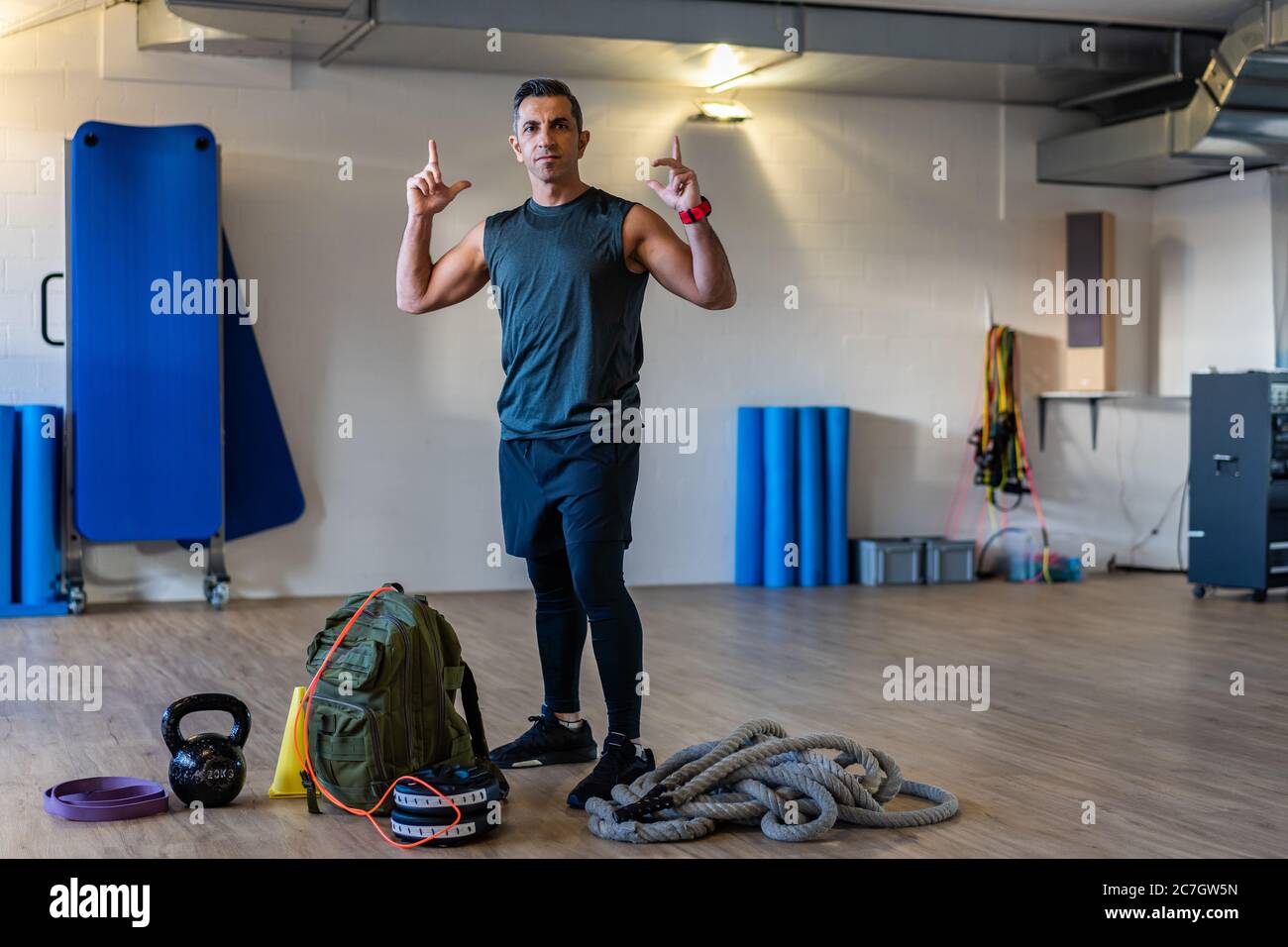 Der Trainer zeigt mit dem Zeigefinger auf die Kamera. Portrait von Boot Camp Instructor steht mit Hanteln, Seil, Sandsack auf Holzboden in Turnhalle. Stockfoto