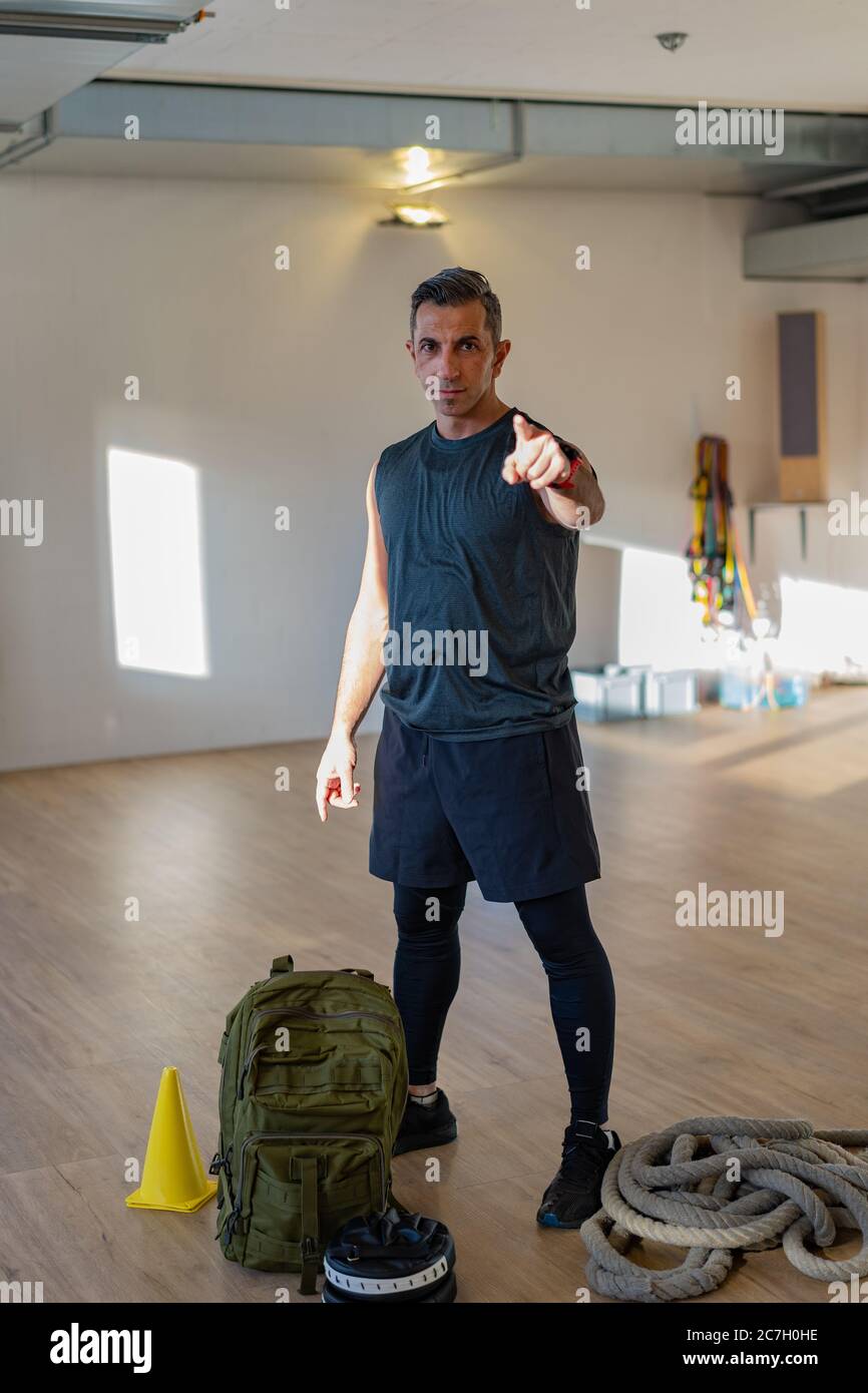 Der Trainer zeigt mit dem Zeigefinger auf die Kamera. Motivierter Boot Camp Instructor steht mit Hanteln, Seil, Sandsack auf Holzboden in der Turnhalle. Stockfoto