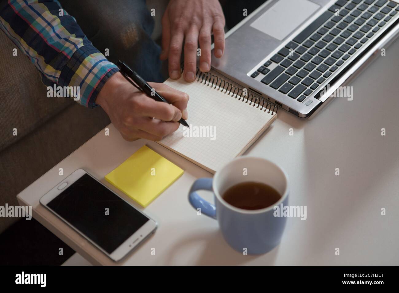 Junger Mann macht Bekanntmachungen in Notebook. Kreativer Freiberufler, der zu Hause an seinem Arbeitsplatz sitzt. Nahaufnahme. Draufsicht Stockfoto