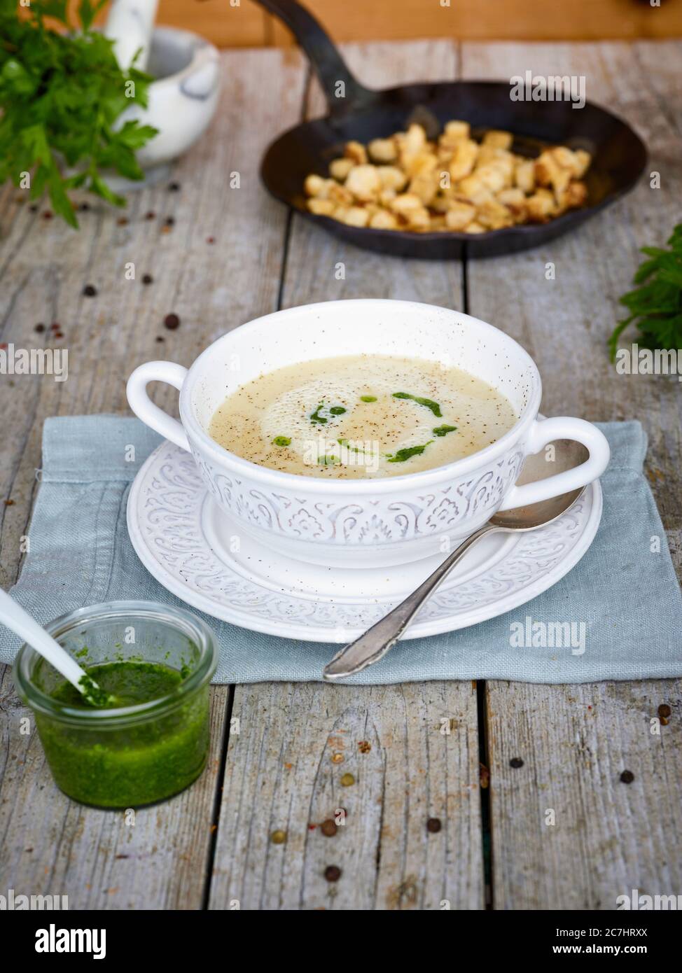 Weincremesuppe in Suppenbecher, Petersilienpesto im Glasgefäß, handgeschmiedete Eisenkanne mit Croutons, Tisch mit unbehandelter Tischplatte Stockfoto