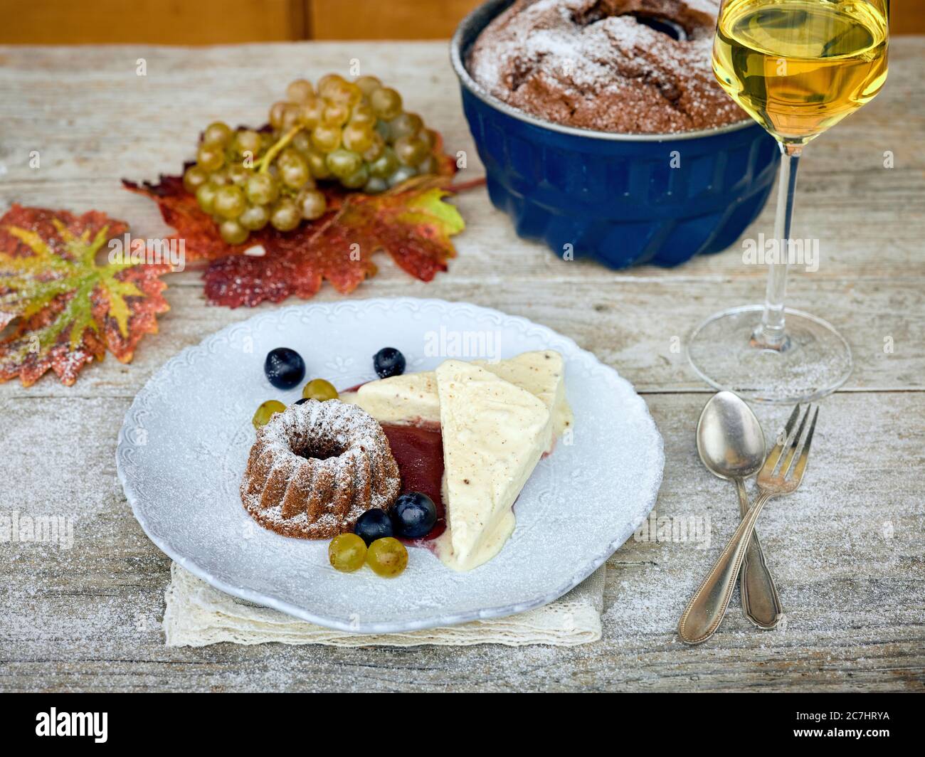 Süßer Weinparfait mit einem kleinen Guglhupf und Weinbeeren auf einem weißen Teller, Weißweinglas gefüllt und Backform, Tisch mit unbehandelter Tischplatte Stockfoto