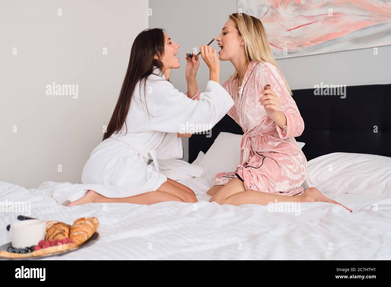 Junge schöne Frau in weißem Bademantel und hübsches Mädchen in rosa Samtmantel Make-up zu einander mit leckeren Frühstück in der Nähe auf einem großen Bett in Stockfoto