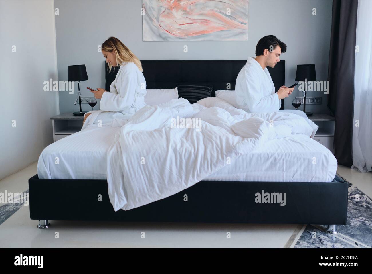 Junge nachdenkliche Brünette Mann und attraktive blonde Frau in weißen Bademänteln mit Handys sitzen auf verschiedenen Seiten des Bettes in modernen Hotel Stockfoto