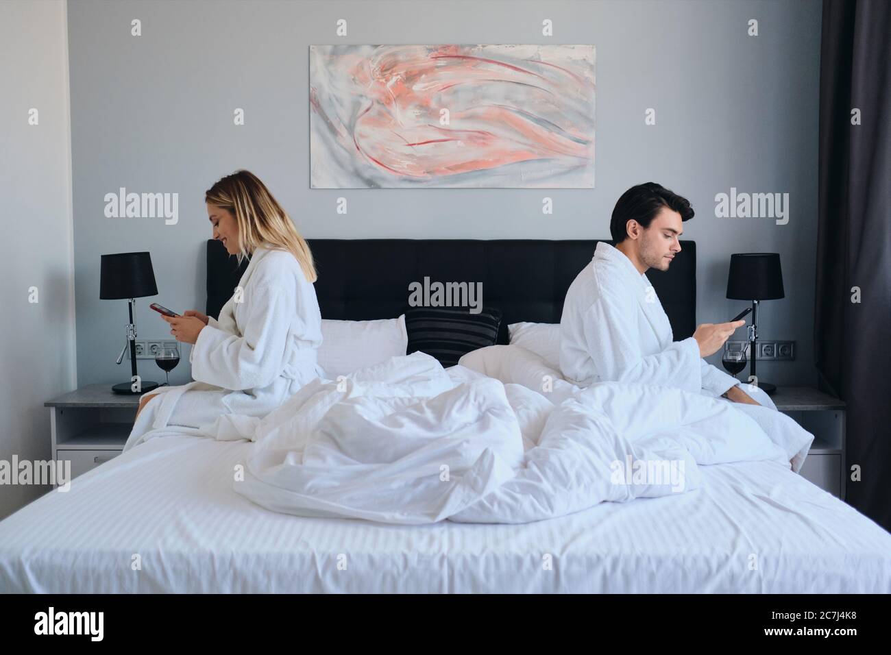 Junge Brünette Mann und hübsche blonde Frau in weißen Bademänteln mit Handys sitzen auf verschiedenen Seiten des Bettes in modernen Hotel Stockfoto