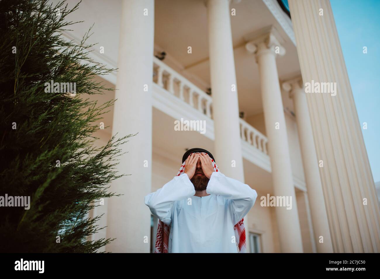 Schöner Mann aus dem Nahen Osten, arabischer muslim in traditioneller Kleidung, der vor dem weißen, schönen, modernen Haus mit den Händen über seinem Gesicht steht Stockfoto