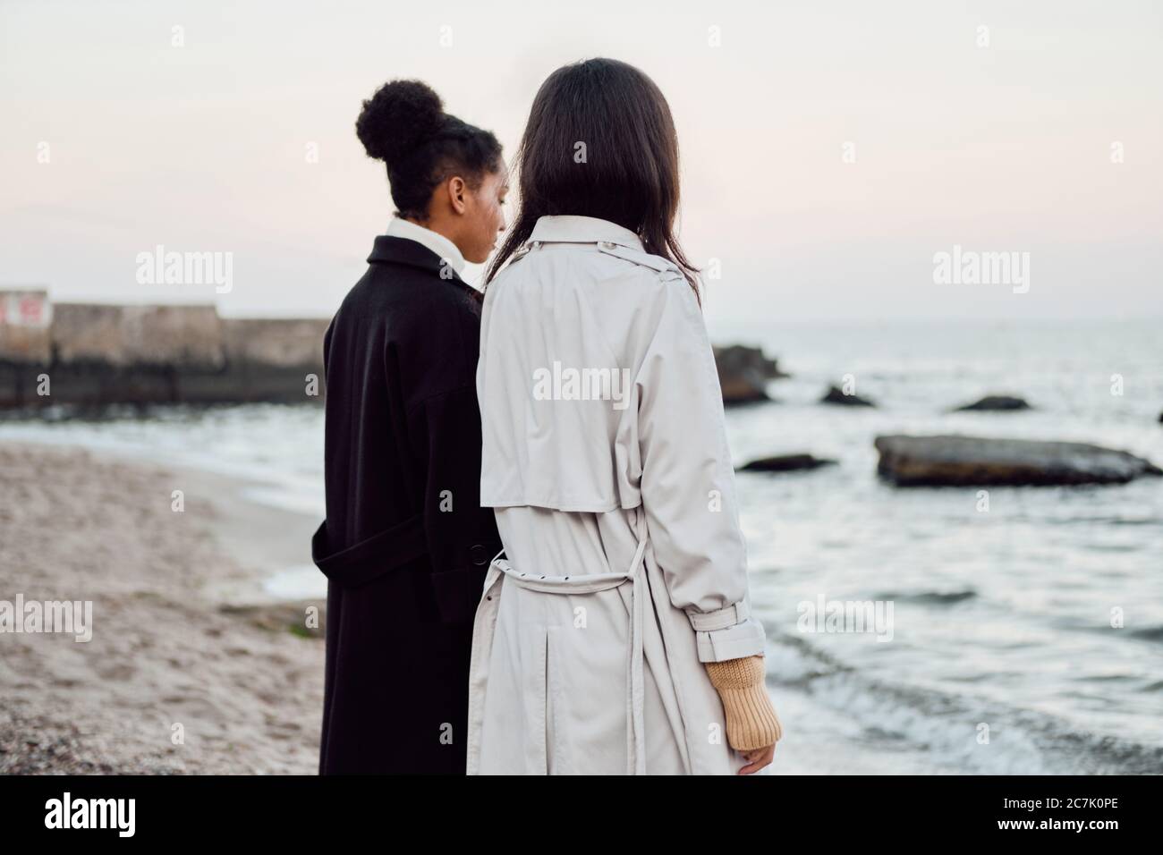 Rückansicht von zwei stilvollen Mädchen in Trenchcoats traumhaft am Meer stehen Stockfoto