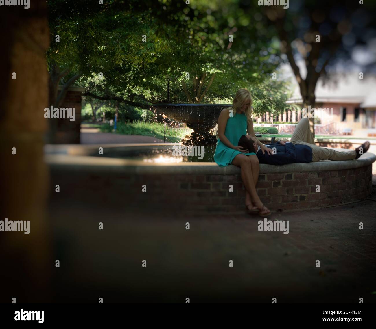 Frau, die neben einem Brunnen sitzt und auf dem ein Mann liegt Ihre Runden in einem Park unter dem Sonnenlicht Stockfoto