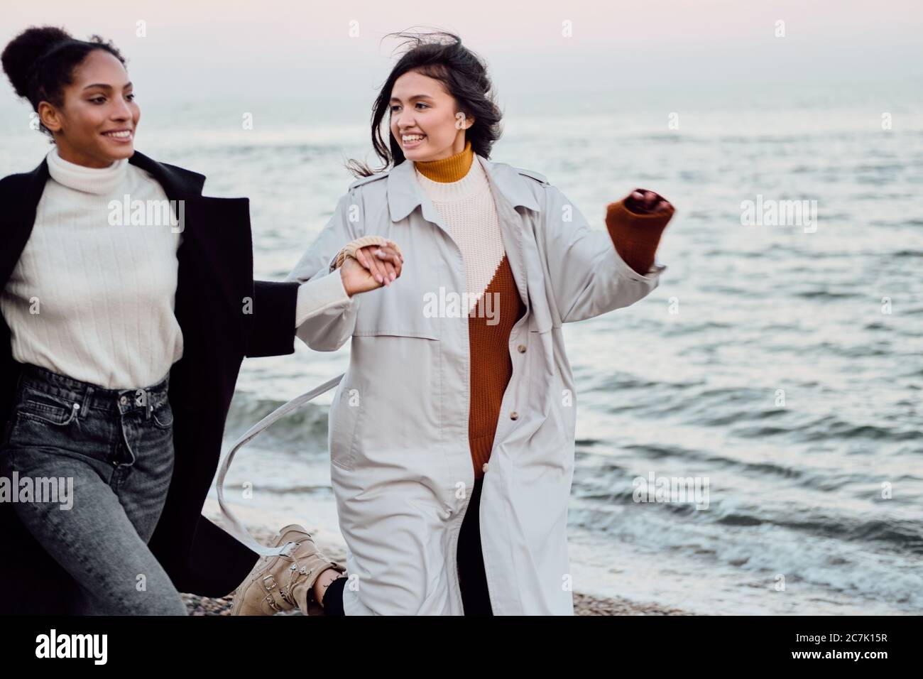 Zwei hübsche stilvolle Mädchen in Trenchcoats freudig am Meer laufen Stockfoto