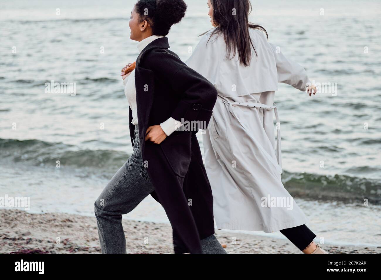 Zwei attraktive Mädchen in Trenchcoats, die von hinten am Meer laufen Stockfoto