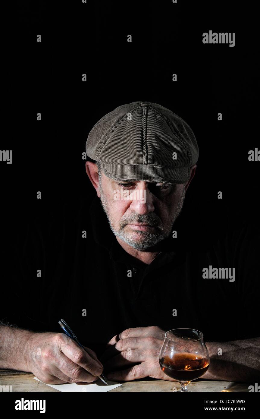 Ein älterer Mann mit Mütze schreibt auf einer Postkarte am Tisch Stockfoto