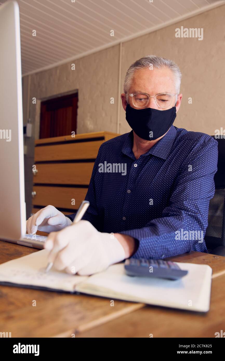 Geschäftsmann, der am Schreibtisch arbeitet und eine Gesichtsmaske vor dem Computer trägt. Stockfoto
