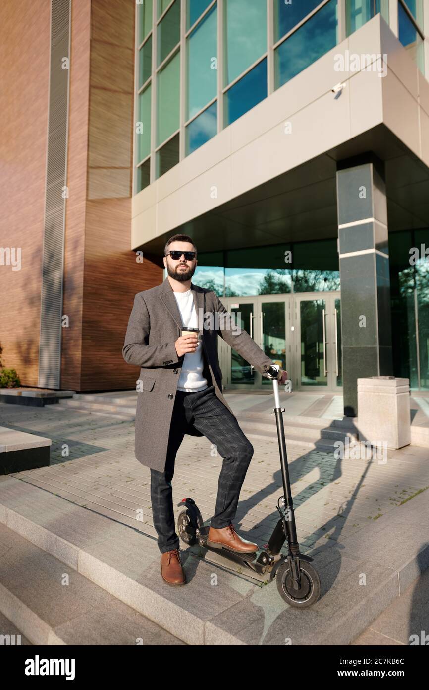 Junge elegante Mann in smart Casualwear mit Kaffee im Freien, während auf Elektroroller von moderner Architektur stehen Stockfoto