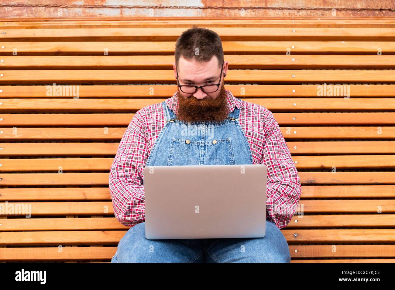 Nahaufnahme Porträt von schweren brutalen bärtigen Hipster Mann in blauen Overalls, kariertes Hemd arbeiten auf Laptop sitzen auf Bank im Freien. Distanzauftrag Stockfoto