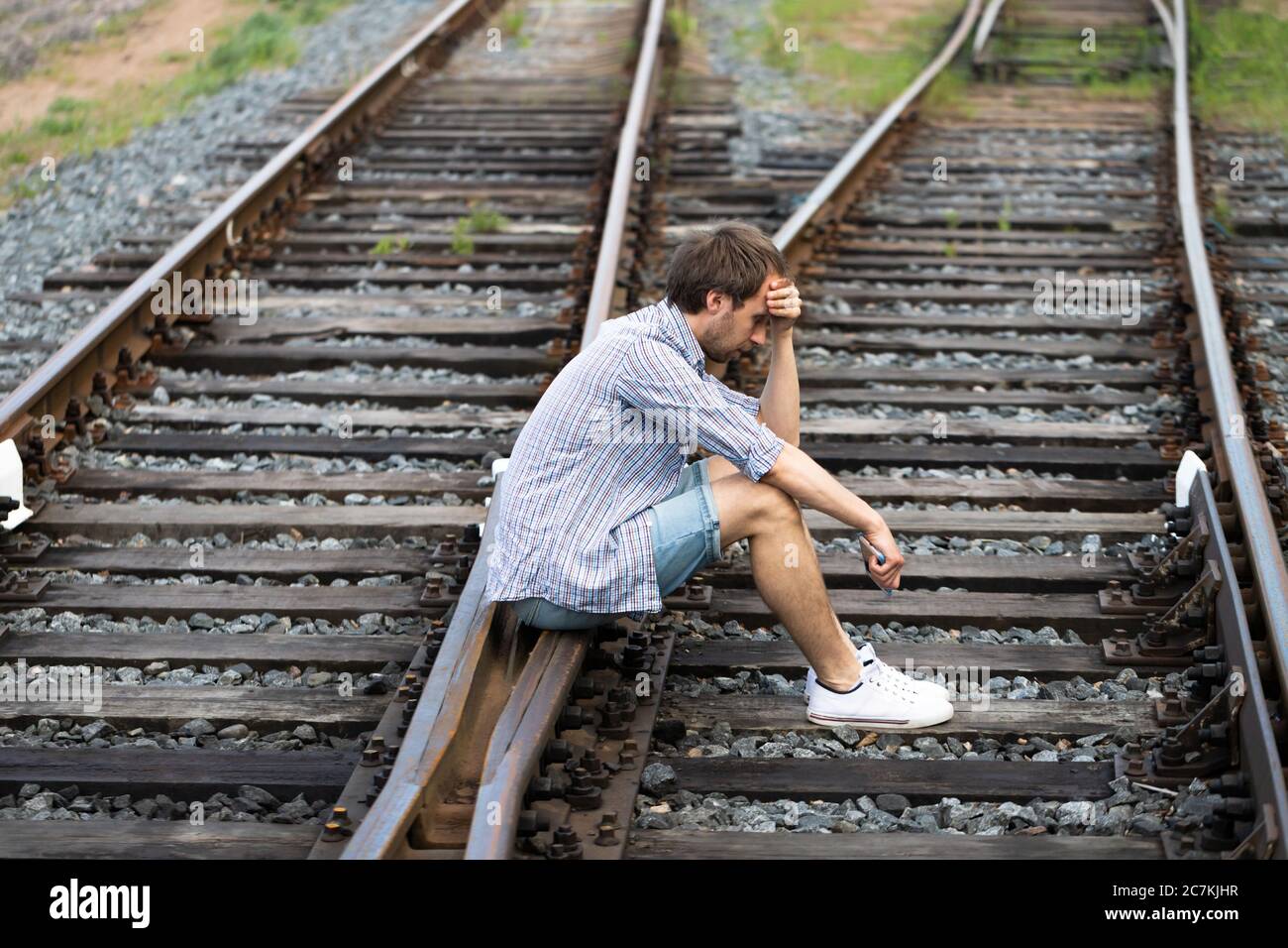 Deprimierter Mann, der auf den Gleisen sitzt, Telefon hält, trifft eine schwierige Entscheidung, in der Vergangenheit zu leben oder seine Zukunft zu ändern. Ändern des Lebenswegs c Stockfoto