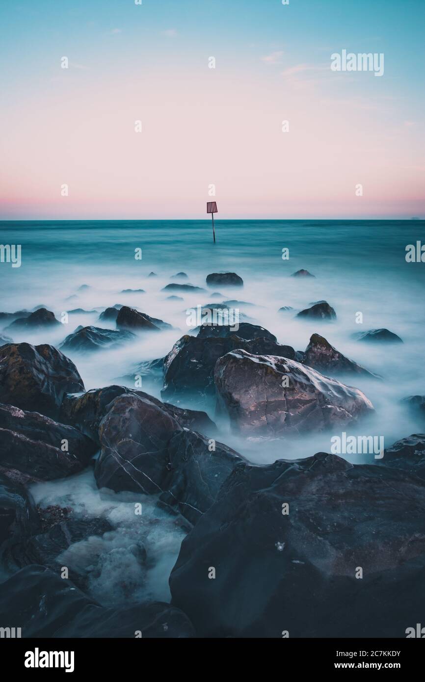 Wellen krachen über Felsen am Meer mit einem Strandmarker im Meer in der Ferne Stockfoto